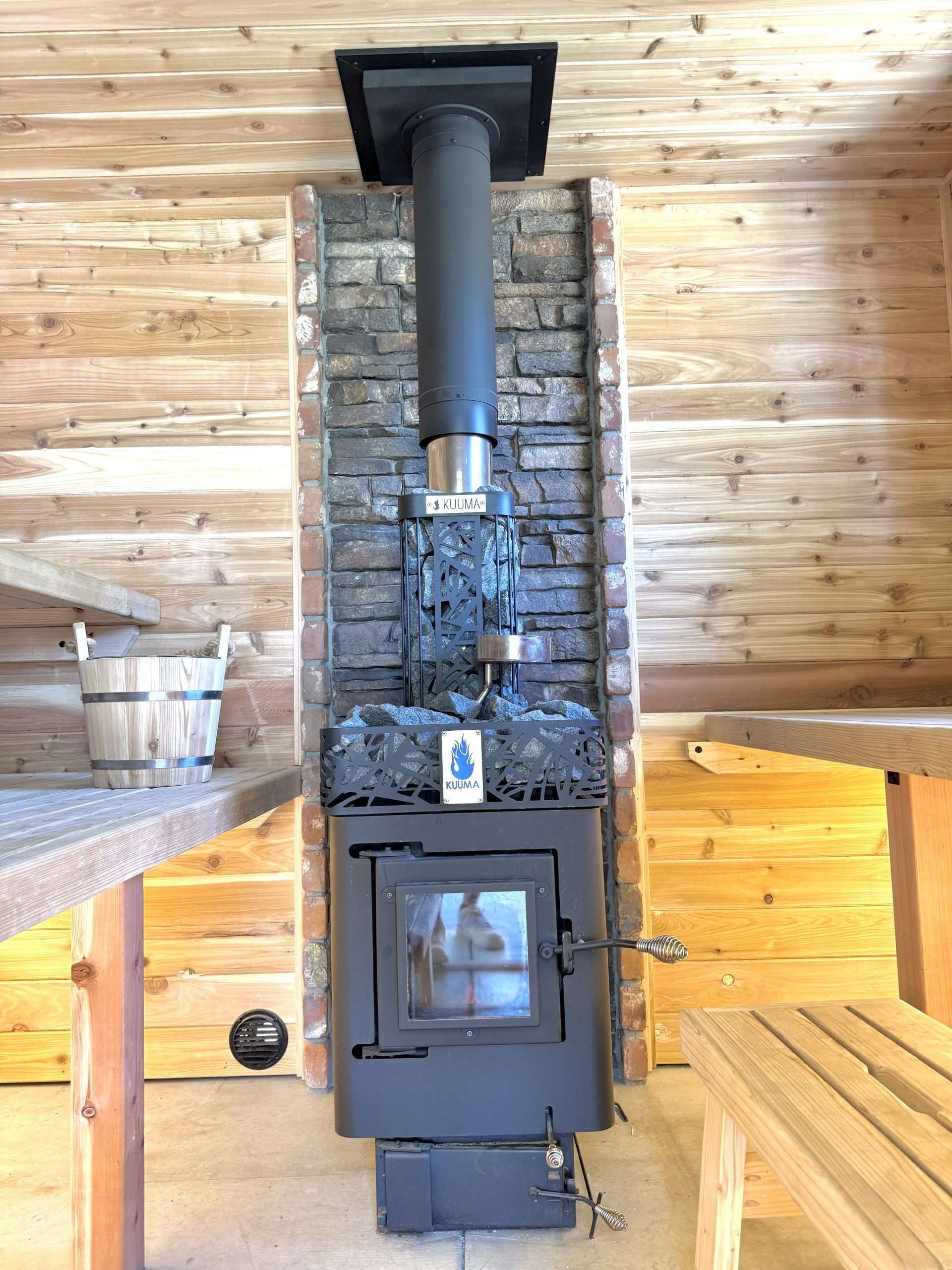Small Kuuma BluFlame wood sauna stove installed in a cedar sauna with a stone wall heat shield behind the stove.